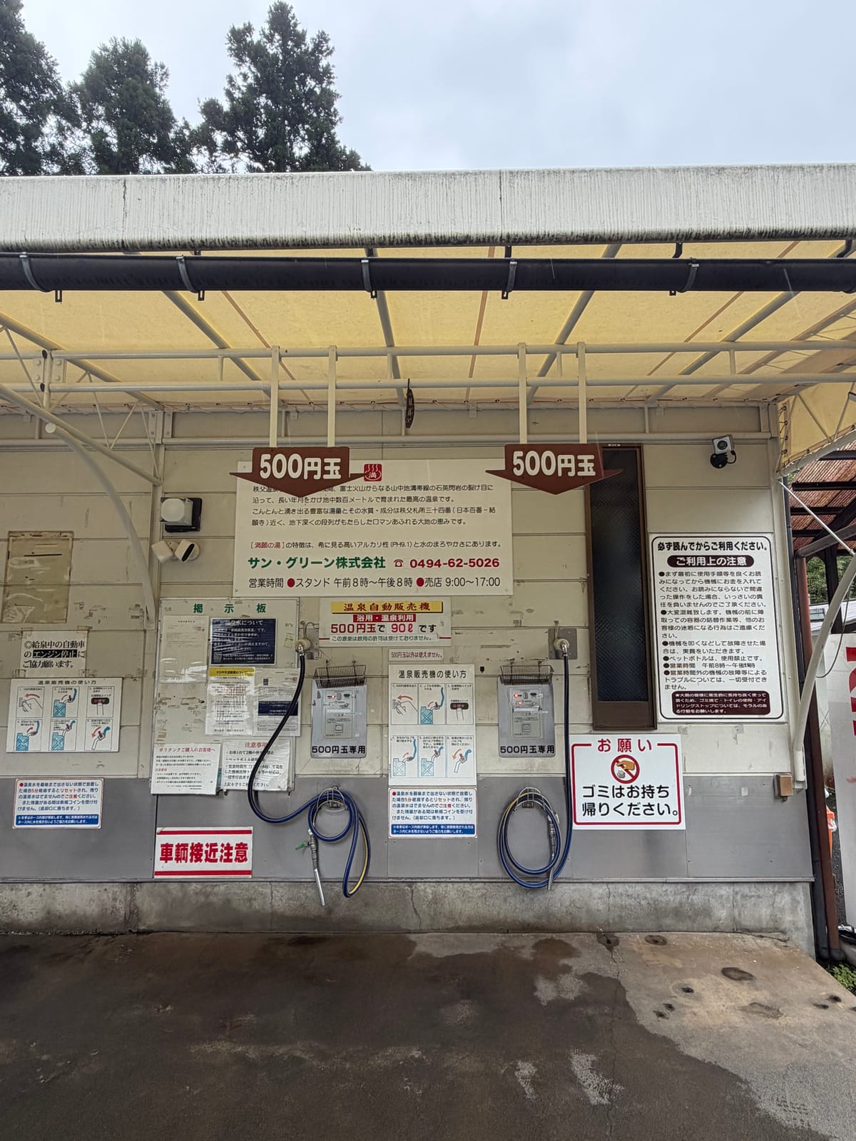 満願の湯源泉所の温泉スタンド全景。屋根の下の壁に500円玉専用の自動販売機が2基並び、それぞれにホースが掛かっている